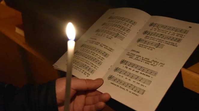 Ruka drží hořící svíčku a otevřený zpěvník s notami